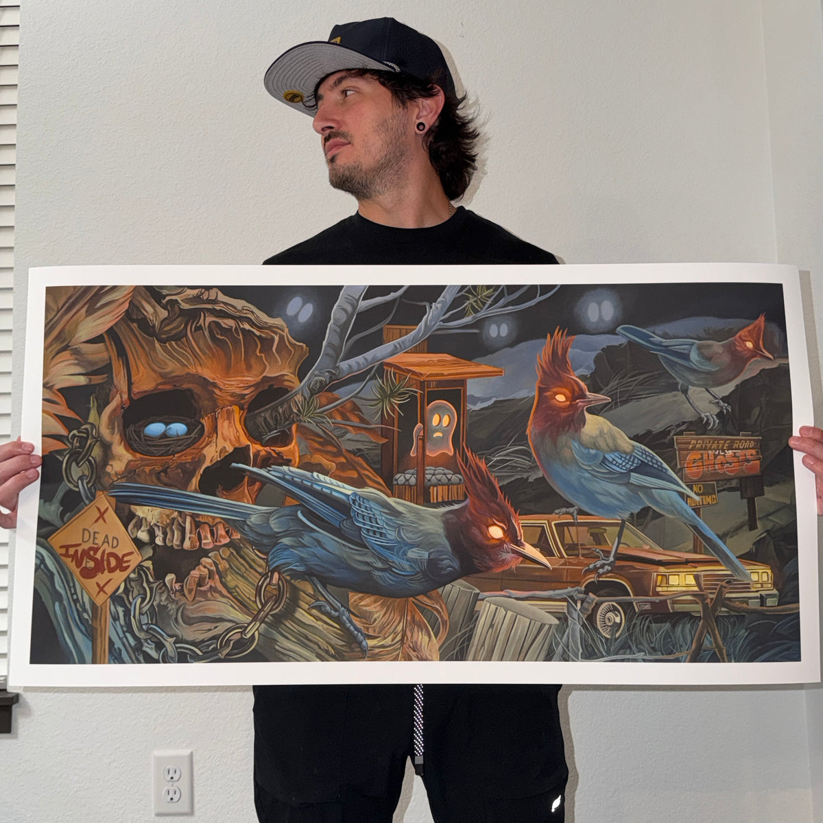 A man wearing a black cap and black clothing holds up a large fine art print. The artwork features a dark, surreal scene with a large skull, a scared ghost, glowing blue birds, an old brown car, and a sign reading "Dead Inside"