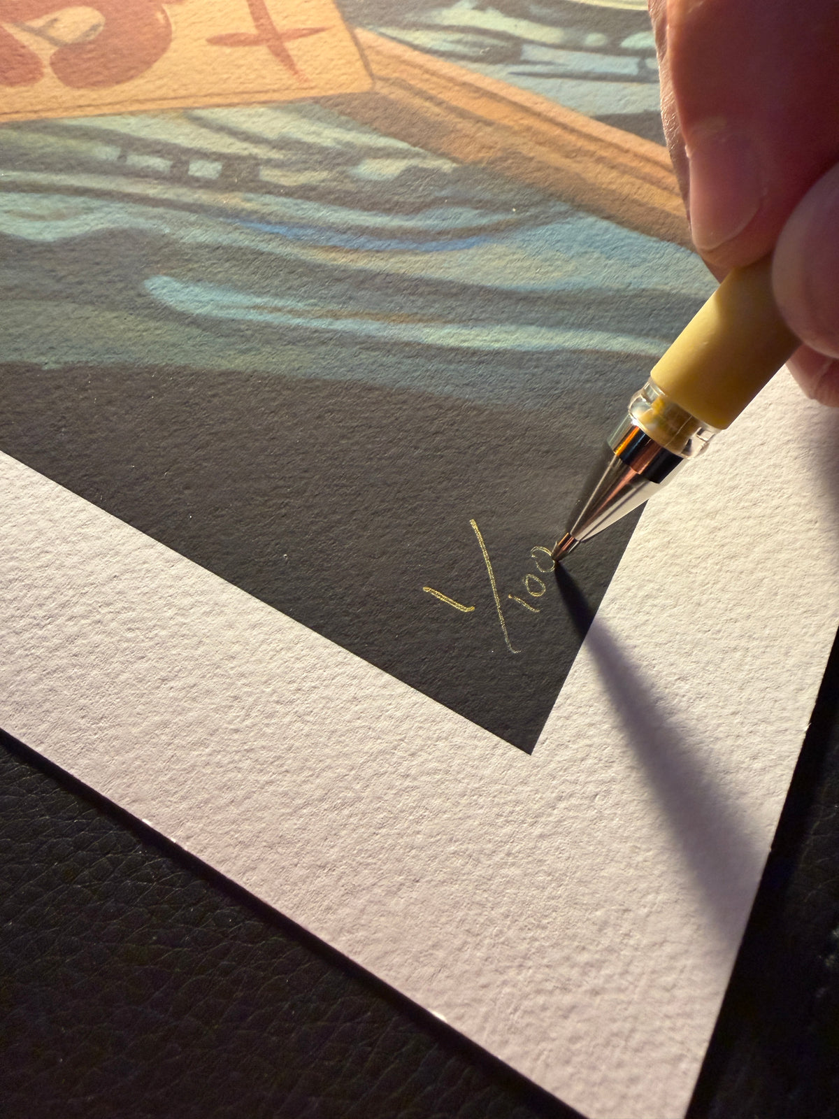 A close-up of a hand holding a yellow and clear fountain pen, writing gold ink signatures on a black and white textured art print.